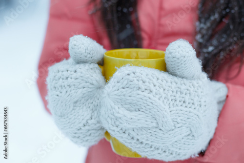 Closeup of female hands in light knitted gloves hold a yellow ceramic cup with a warming drink, tea or coffee outside on winter snow day. Winter and Christmas time concept. Space for text