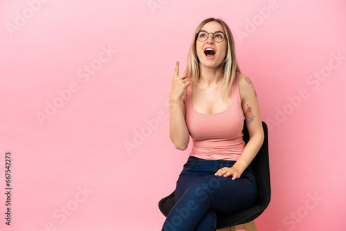 Young woman sitting on a chair over isolated pink background pointing up and surprised