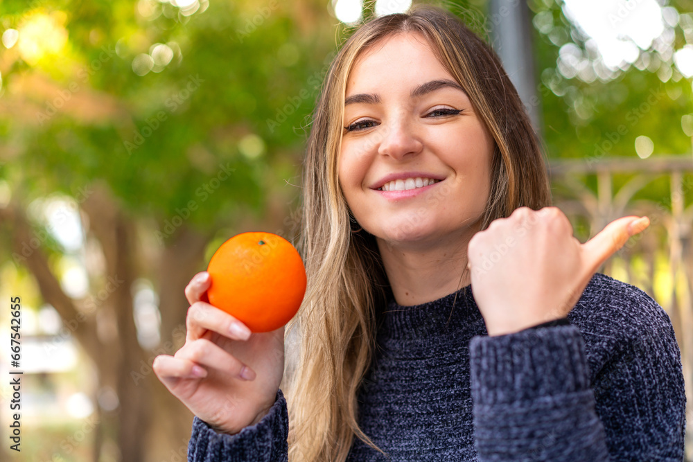 Young pretty Romanian woman holding an orange at outdoors pointing to the side to present a product Young pretty Romanian woman holding an orange at outdoors pointing to the side to present a product