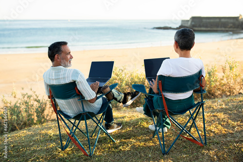 Senior spouses sit together with laptops at scenic ocean viewpoint