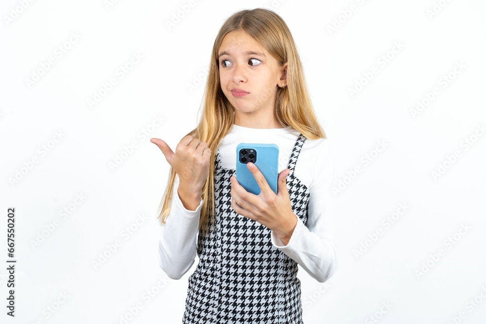 Beautifu blonde kid girl points thumb away and shows blank space aside, holds mobile phone for sending text messages. Beautifu blonde kid girl points thumb away and shows blank space aside, holds mobile phone for sending text messages.