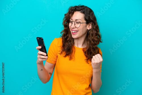 Young caucasian woman isolated on blue background using mobile phone and doing victory gesture