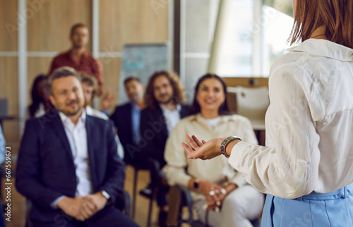 Woman standing back speaking on a business training in office during a conference or seminar for a group of happy people company employees sitting on chairs in a row in meeting room.