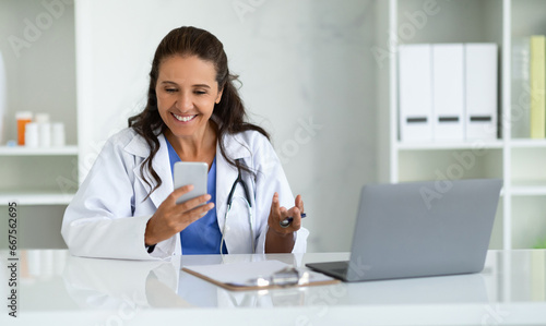 Cheerful mature lady doctor sitting in her cabinet, using phone