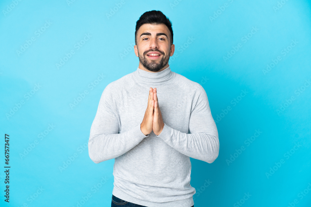 Caucasian man over isolated blue background keeps palm together. Person asks for something Caucasian man over isolated blue background keeps palm together. Person asks for something
