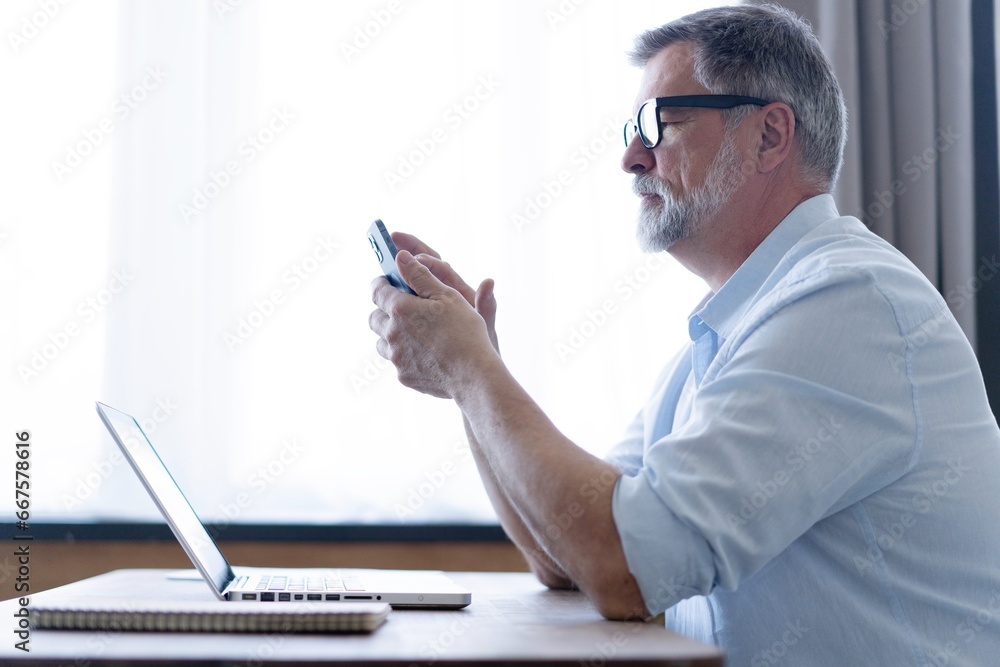 Handsome smiling senior man wearing glasses using mobile phone while sitting at his workplace with laptop at home Handsome smiling senior man wearing glasses using mobile phone while sitting at his workplace with laptop at home