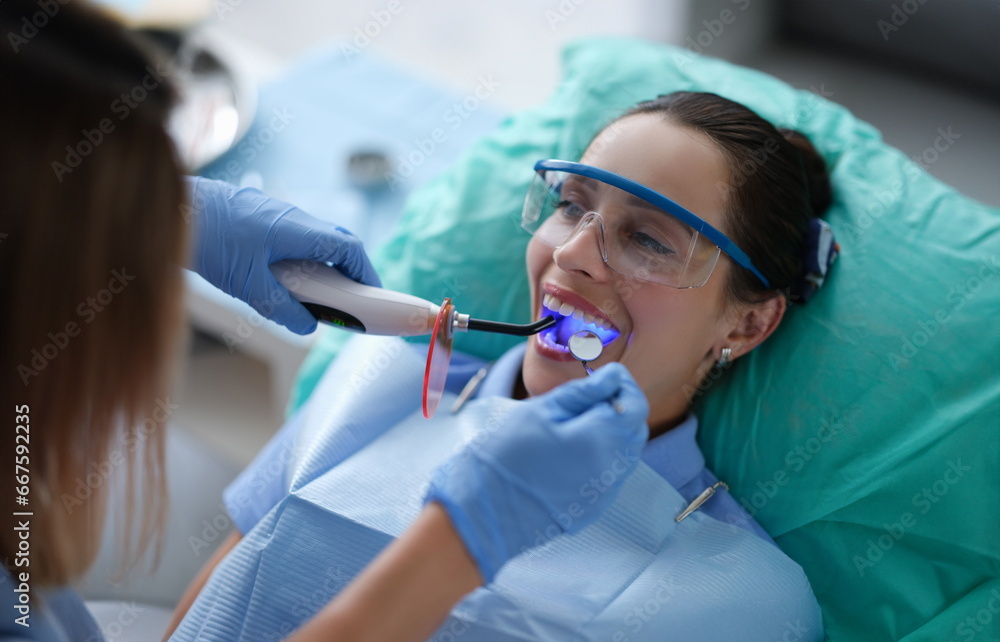 Young woman dentist in protective gloves checking dental seal, fixing photopolymer lamp during stomatology procedure. Dentist specialist visiting patient in dental clinic.