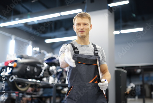 Young smiling master man in uniform stretches his arms forward in car service. High-quality service of motorcycles and cars concept