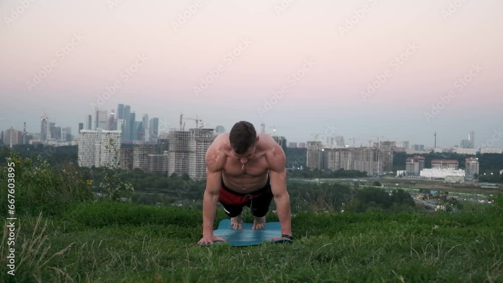A muscular guy practices yoga asanas, stretching alone in a green meadow. Focused topless man doing an evening workout against the backdrop of a big city. Sports yogi meditates on a fitness mat. A muscular guy practices yoga asanas, stretching alone in a green meadow. Focused topless man doing an evening workout against the backdrop of a big city. Sports yogi meditates on a fitness mat.