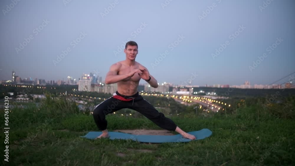 A muscular guy practices yoga asanas, stretching alone in a green meadow. Focused topless man doing an evening workout against the backdrop of a big city. Sports yogi meditates on a fitness mat A muscular guy practices yoga asanas, stretching alone in a green meadow. Focused topless man doing an evening workout against the backdrop of a big city. Sports yogi meditates on a fitness mat