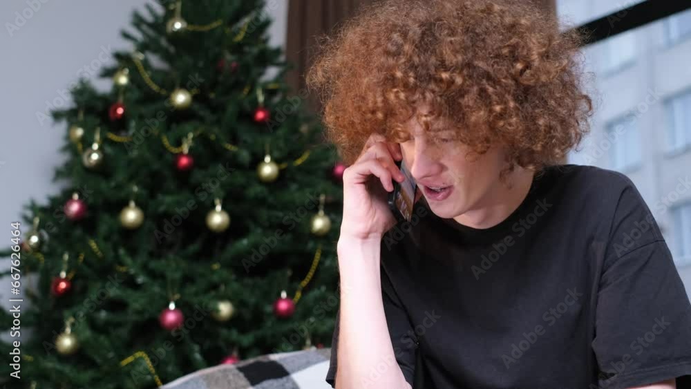 A young man is talking on the phone near a colorful winter tree in a room. Christmas greetings and gifts A young man is talking on the phone near a colorful winter tree in a room. Christmas greetings and gifts