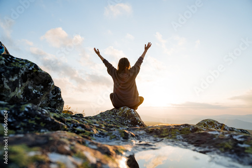 Man joyfully raising his hands up, happy moments in nature, looking at the sun, back view of man sitting on a stone, feeling of freedom, © Aleksey