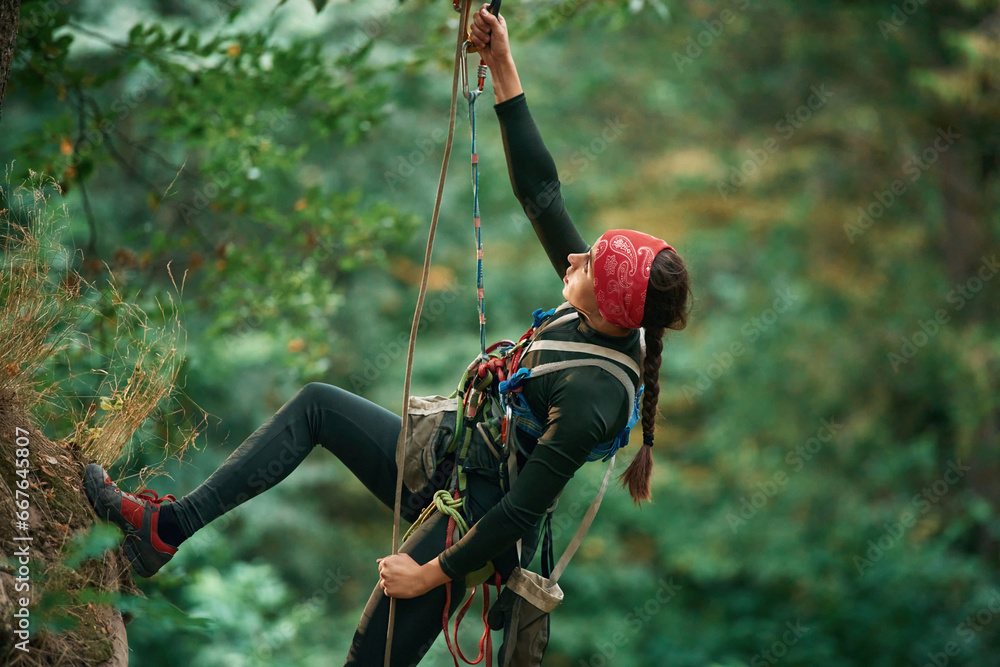 In sportive clothes, high up on the rope. Woman is doing climbing in the forest by the use of safety equipment - obrazy, fototapety, plakaty 