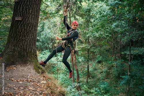 Looking to the side, hanging on the rope. Woman is doing climbing in the forest by the use of safety equipment