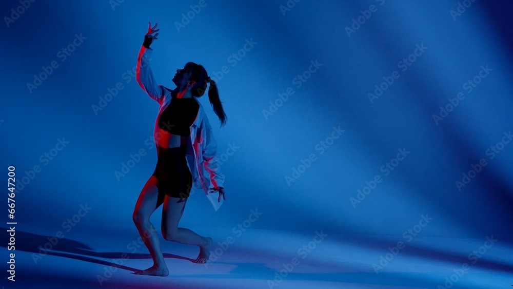 Young woman wearing a top, shorts and a shirt performing contemporary dance in studio. Neon blue and red color scheme, shadowed background. Full length.