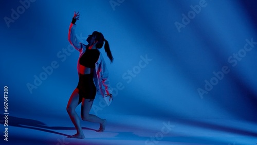 Fototapeta Naklejka Na Ścianę i Meble -  Young woman wearing a top, shorts and a shirt performing contemporary dance in studio. Neon blue and red color scheme, shadowed background. Full length.