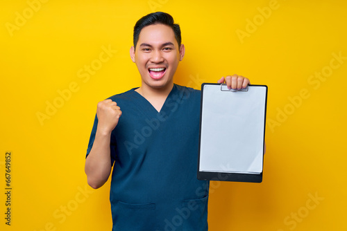 Excited professional young Asian male doctor or nurse wearing a blue uniform holding clipboard with blank paper and celebrating success isolated on yellow background. Healthcare medicine concept photo