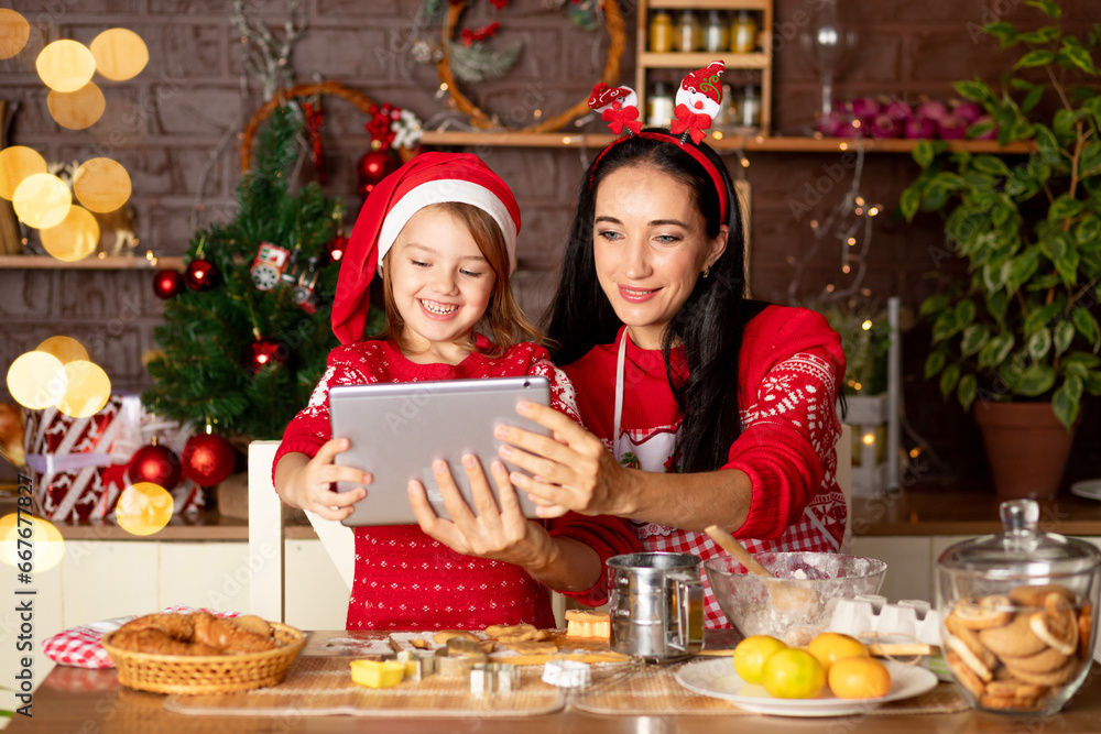 mom and daughter are talking via video link, passing greetings on a tablet in a dark kitchen with a Christmas tree for New Year or Christmas, smiling and having fun together in a Santa Claus hat