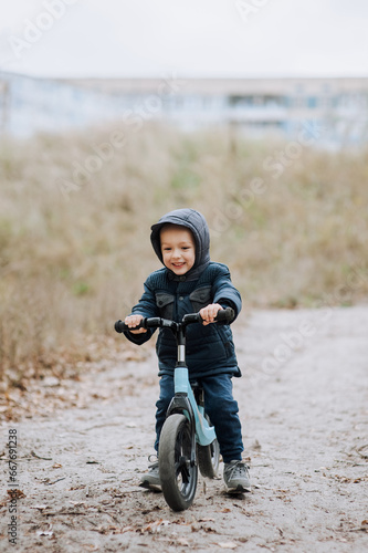 Happy smiling handsome child, preschool boy rides fast on a blue balance bike, bicycle without wheels on a road with a slope outdoors in autumn. Photography, portrait, childhood concept, sports. © shchus