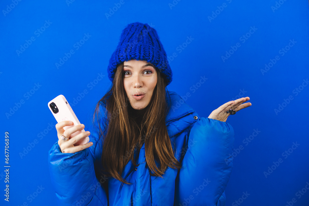 Shocked brunette woman in blue puffer jacket hold mobile phone isolated on blue background studio. Shocked woman with open mouth, hand near face wow win emotions. Amazing useful app. Shocked brunette woman in blue puffer jacket hold mobile phone isolated on blue background studio. Shocked woman with open mouth, hand near face wow win emotions. Amazing useful app.