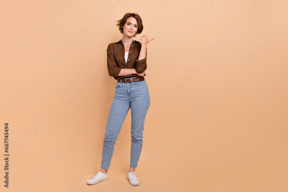 Full length photo of cute shiny girl dressed brown shirt pointing thumb empty space isolated beige color background Full length photo of cute shiny girl dressed brown shirt pointing thumb empty space isolated beige color background