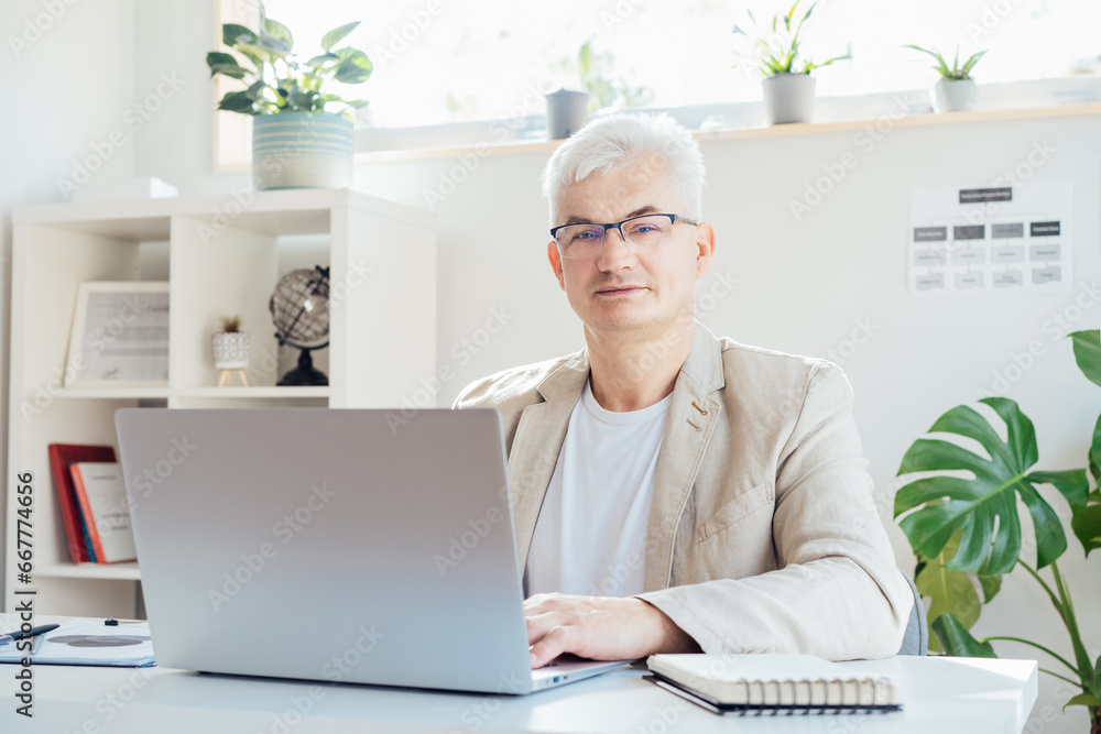 Portrait of smiling grey hair middle-aged man working on laptop while sitting at his work place in office. Confident, experienced senior male professional. Small business entrepreneur manage business Portrait of smiling grey hair middle-aged man working on laptop while sitting at his work place in office. Confident, experienced senior male professional. Small business entrepreneur manage business