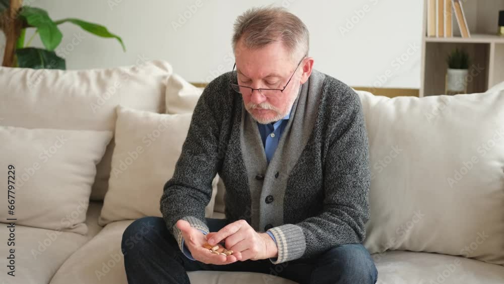 Middle aged senior man holding medical pills sitting on couch at home. Mature old senior grandfather taking medication cure pill vitamin. Age prescription medicine healthcare therapy concept Middle aged senior man holding medical pills sitting on couch at home. Mature old senior grandfather taking medication cure pill vitamin. Age prescription medicine healthcare therapy concept