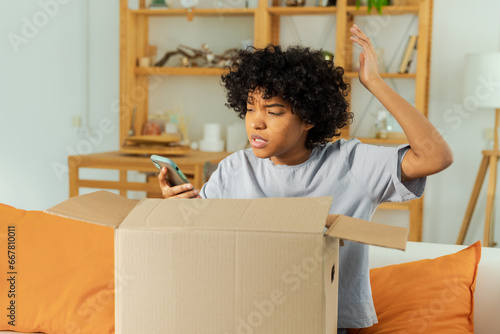 Unhappy angry african woman unpack delivered box receiving damaged shipping mistake problem in postal service. Annoyed confused customer client receiving wrong parcel checking online app on smartphone © Юлия Завалишина