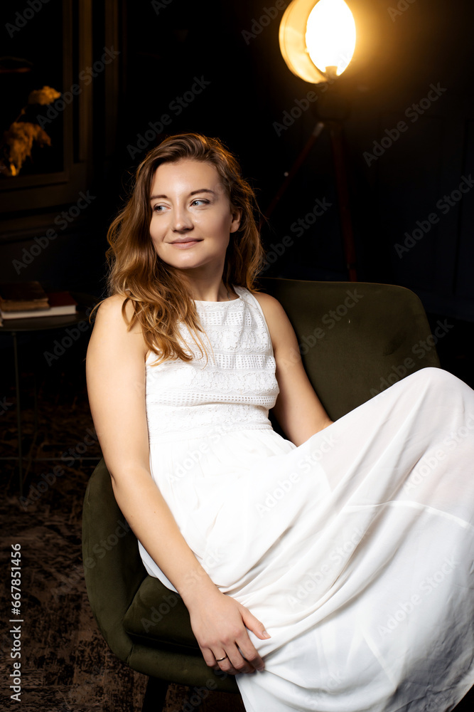 In a shadowy space, a young lady dons a white gown, sitting gracefully on a chair. Her long locks frame her face as she directs a smiling glance elsewhere In a shadowy space, a young lady dons a white gown, sitting gracefully on a chair. Her long locks frame her face as she directs a smiling glance elsewhere