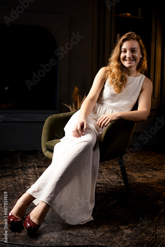 In the shadows, a young woman, her hair flowing, rests on a chair, casting a smile towards the camera lens in the darkness of the room