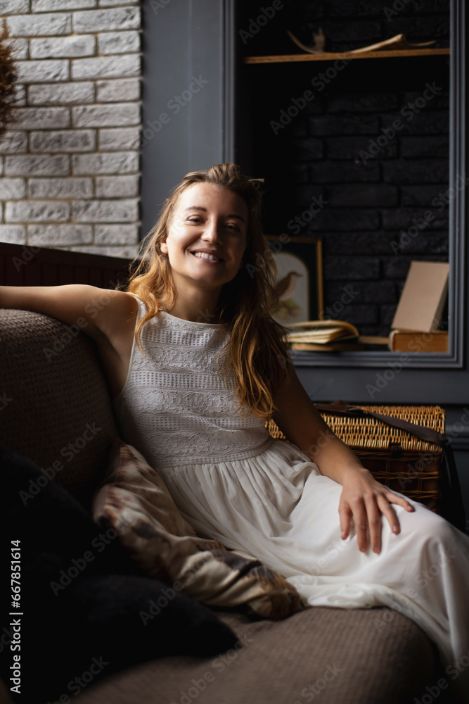 A young woman in a long white dress with long hair sits on the sofa smiling and looking at the camera. concept, have a good time A young woman in a long white dress with long hair sits on the sofa smiling and looking at the camera. concept, have a good time