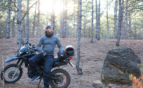 caucasian man with a helmet and gloves sitting near a motorbike in the forest at sunset light