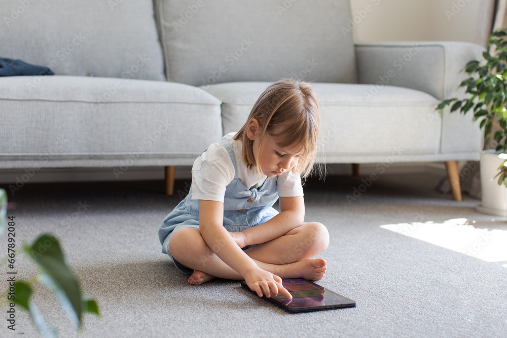 A cute girl looks at the tablet screen while sitting on the floor in the room. Children's educational apps for tablet. 