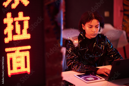 Girl typing on laptop keyboard in bar with hieroglyphs meaning best ramen while sitting by table in cyberpunk cafe lit with neon lights