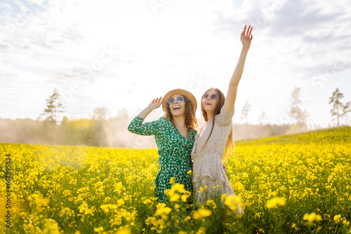 Two cheerful girlfriends have a great summer time walking through a blooming rapeseed field. The concept of walking, active lifestyle, holidays.