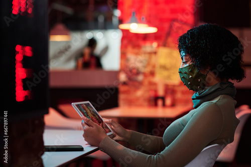 Side view of young woman in facial mask chilling in cyberpunk cafe and looking at tablet screen while watching online video or playing game
