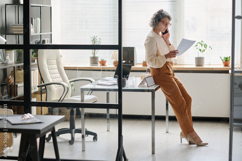 Side view of mature professional auditor or broker looking through contract terms or financial paper and talking on mobile phone Side view of mature professional auditor or broker looking through contract terms or financial paper and talking on mobile phone