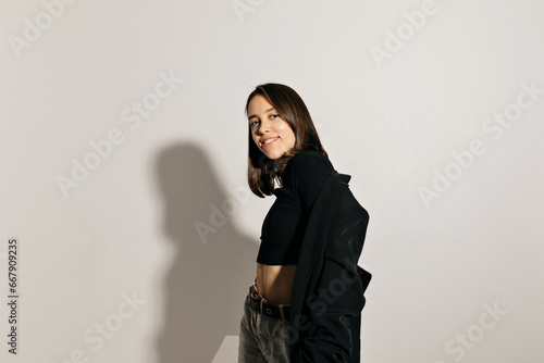 Beautiful young caucasian girl modestly laughs standing on white background. Model with dark hair wears black jacket. Lifestyle concept, female beauty concept