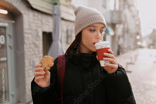 Interested brown-haired woman in beige cap holding cup of cappuccino and cookies and enjoying coffee break in the city.  Photo of enchanting confident girl drinking coffee on the road.