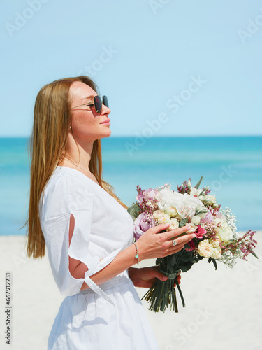 Obraz na płótnie Joyful Young Woman Holding a Bouquet of Flowers on a Beach with Blue Sky Background w salonie