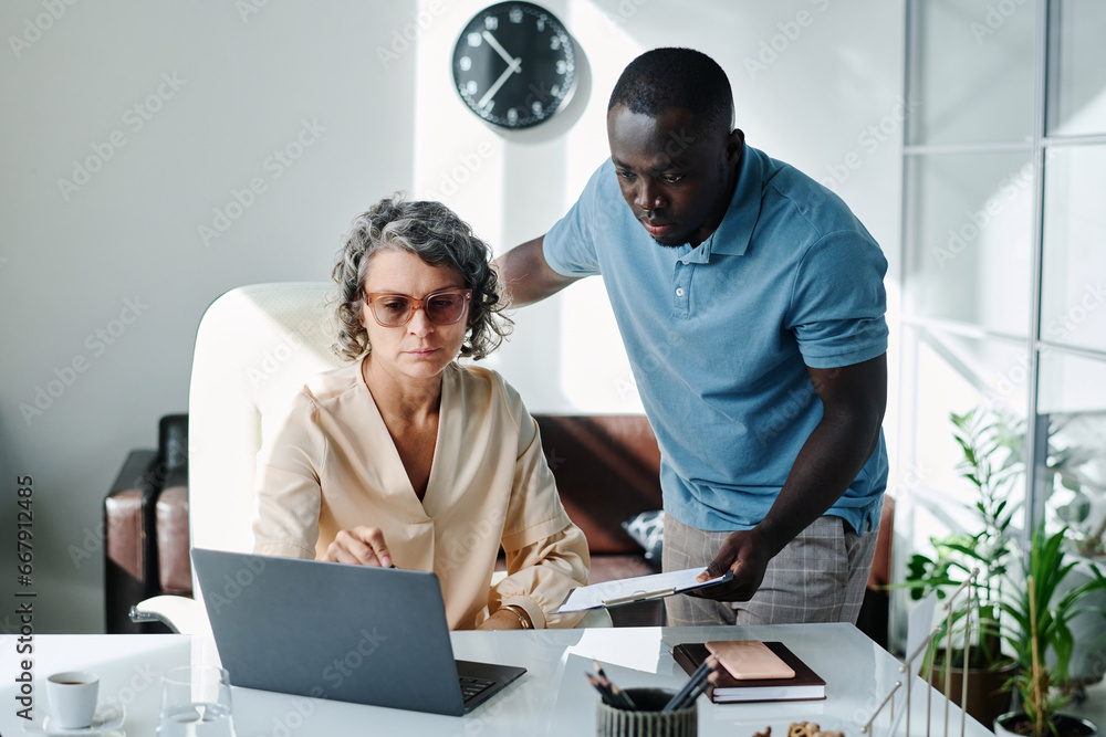 Confident female director of company pointing at laptop screen while explaining online data to young African American male subordinate Confident female director of company pointing at laptop screen while explaining online data to young African American male subordinate