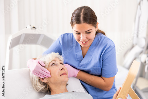 Satisfied senior woman looking at mirror after professional cosmetology treatment with beautician in modern beauty salon indoors