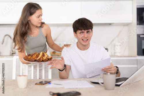 Engaged young boy working on laptop on kitchen-table and his wife bringing tart in the plate