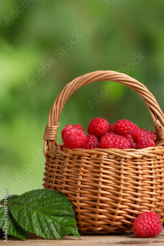 Wicker basket with tasty ripe raspberries and green leaves on table against blurred background, closeup