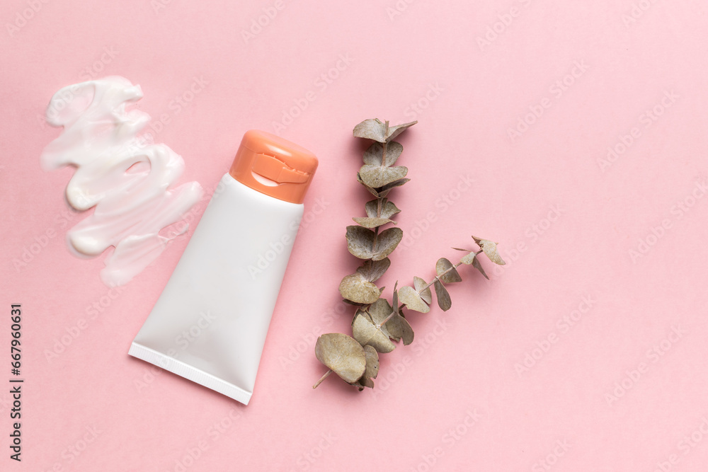 A white tube, smeared cream and a sprig of dried eucalyptus on a pink background. The concept of body care. A white tube, smeared cream and a sprig of dried eucalyptus on a pink background. The concept of body care.