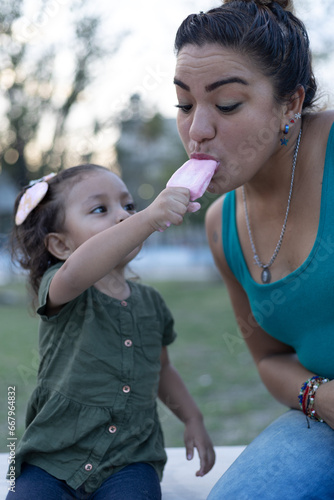 mexican mother and daughter eating snow popsicle in park