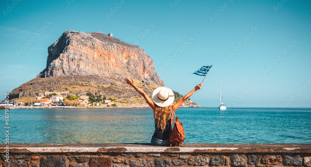 Obraz premium Happy woman tourist with bag and flag enjoying vacation in Greece- Monemvasia island