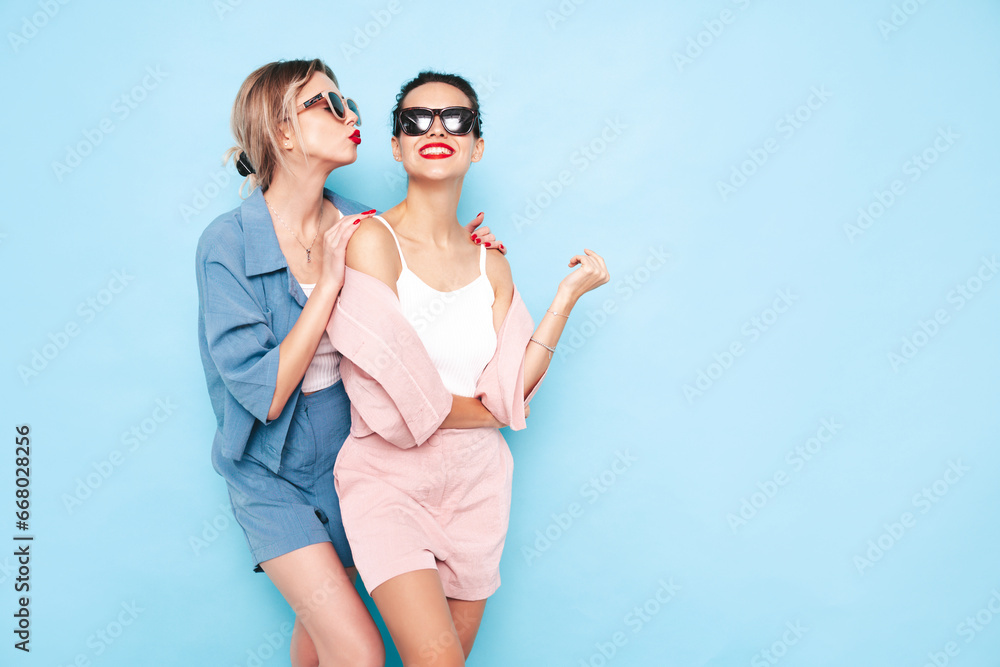 Two young beautiful smiling brunette hipster female in trendy summer clothes. Sexy carefree women posing near blue wall in studio. Positive models having fun. Cheerful and happy. In sunglasses