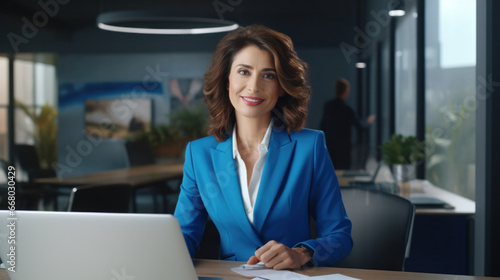 Radiant CEO: Middle-Aged Businesswoman in Blue Suit Skillfully Maneuvers Laptop, Epitomizing Professionalism and Leadership from Her Office Desk.