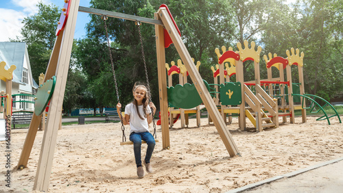 The child plays in the outdoor playground. Active little girl swings on a colorful swing. Healthy summer activity for kids.